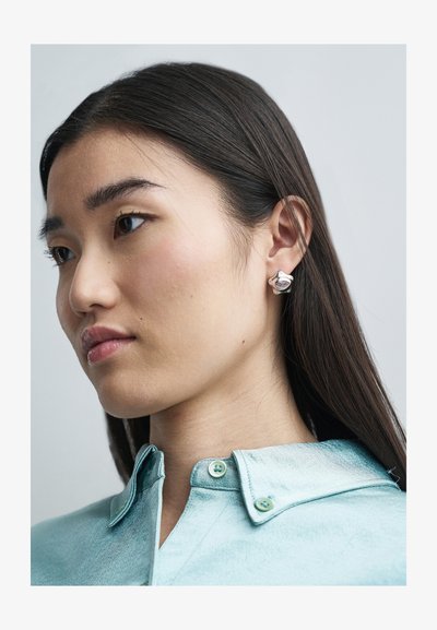 Boucles d'oreilles en argent avec un design arrondi et des accents texturés. Le modèle porte une chemise en satin bleu clair avec un col et des boutons verts.