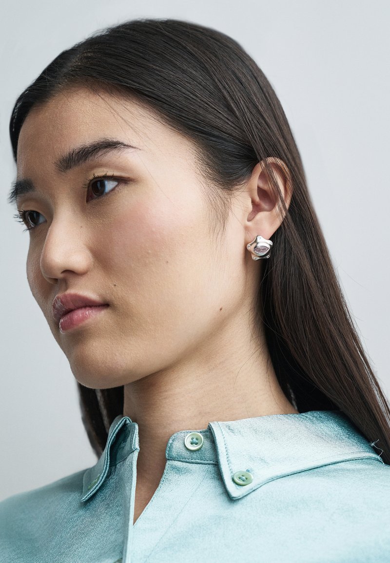 Silver earrings with a rounded design and textured accents. The model wears a light blue satin shirt with a collar and green buttons.