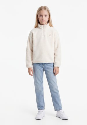 Jeune fille aux longs cheveux blonds portant un pull en polaire crème, un jean bleu clair et des baskets blanches, debout devant un fond uni.