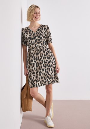 Femme en robe à manches courtes imprimé léopard, baskets blanches, tenant un sac marron, appuyée contre un mur blanc, souriante et regardant vers la gauche.