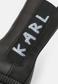 Stivale nero con tomaia in pelle elegante e pannello laterale elastico che presenta la scritta "KARL" in vernice azzurro chiaro su sfondo nero. Suola testurizzata.