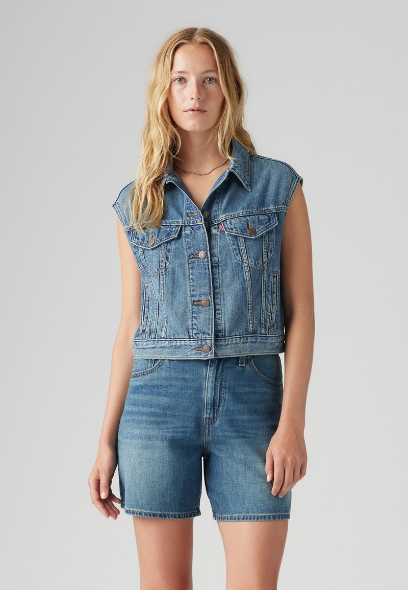 Denim sleeveless jacket in light blue, featuring a button front, two chest pockets, and a cropped cut, paired with matching blue denim shorts.