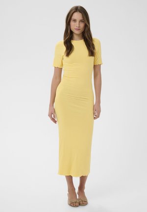 Žena stojaca v priliehavých rebrovaných žltých midi šatách s krátkymi rukávmi a s béžovými otvorenými sandálmi na podpätku, na jednoduchom sivom pozadí.