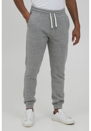 Mand iført lysegrå joggingbukser med hvide snørrebånd og hvide sneakers, stående mod en ensfarvet baggrund.