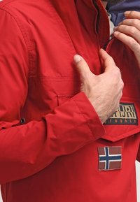 Giacca leggera rossa con chiusura a zip, dotata di una toppa con logo e un emblema della bandiera della Norvegia. Il materiale sembra liscio e resistente.