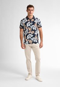 Camisa polo de manga corta en azul marino oscuro con un estampado floral en azul claro y beige. Combinada con pantalones beige y zapatillas blancas.