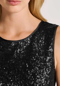 Frau trägt ein schwarzes ärmelloses Top, das mit kleinen glänzenden Pailletten bedeckt ist, und zeigt ihren Hals, ihre Schultern sowie einen Teil ihres Gesichts und Haares.