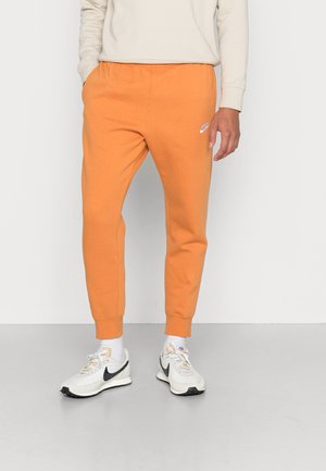 Osoba w pomarańczowych spodniach dresowych Nike, białych skarpetkach i białych butach sportowych Nike, stojąca na tle jasnego, jednolitego tła.