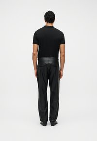 Άνδρας που στέκεται με την πλάτη, φοράει μαύρο κοντομάνικο πουκάμισο και μαύρο δερμάτινο παντελόνι με πίσω τσέπες, σε απλό λευκό φόντο.