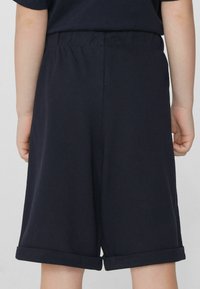 Shorts en coton navy avec une taille élastique et des ourlets retroussés, présentant un tissu texturé. Vue montrant l'arrière avec les mains sur les côtés.