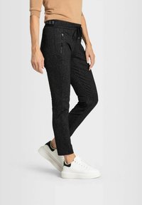 Kvinna som bär svarta strukturerade joggers med dragkedjefickor, beige topp och vita sneakers med svarta detaljer mot en enkel bakgrund.