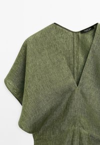 Robe en lin vert texturé avec des manches courtes larges et un profond décolleté en V, montrant le haut du buste et la zone des épaules.