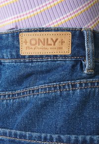 Džínové kalhoty s hnědou koženou etiketu označenou "ONLY Blue Denim anno 1995" s viditelným šitím a klasickou modrou barvou.