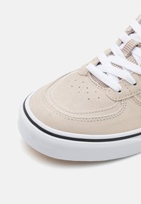 Bout de basket en daim beige avec lacets blancs, perforations et semelle épaisse en caoutchouc blanc avec bordure noire sur fond blanc.
