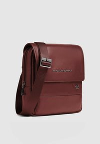 Bolso mensajero de cuero rojo con cierre de solapa, correa ajustable y logo plateado. Textura lisa con un compartimento con cremallera.