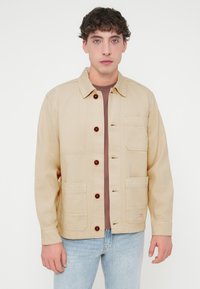 Μπεζ βαμβακερό σακάκι με κούμπωμα μπροστά με κουμπιά, τσέπη στο στήθος, δύο μπροστινές τσέπες και καφέ κουμπιά. Φοριέται πάνω από καφέ πουκάμισο.