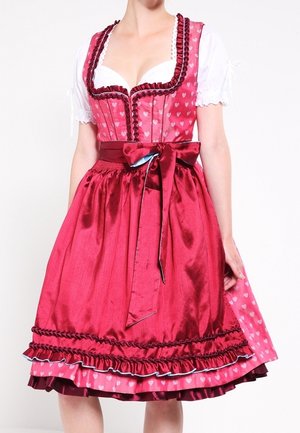 Kvinde iført traditionel rød dirndl kjole med hjerte mønstre, hvid bluse, flæset nederdel og satinsløjfe bundet i siden af taljen.