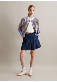 Gestreepte cardigan in blauw en bruin, gecombineerd met een donkerblauwe, geplooide denimrok en beige sneakers. Model poseert met handen in de zakken.