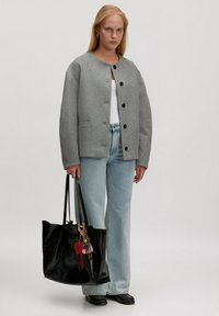 Frau trägt eine graue Knopfjacket, ein weißes Oberteil, hellblaue Jeans, schwarze Schuhe und hält eine große schwarze Tragetasche mit bunten Schlüsselanhängern.