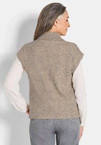 Hellbraunes Strickwesten mit gerippter Textur, hohem Ausschnitt und Raglanärmeln, kombiniert mit einem weißen Langarmshirt und grauen Jeans.