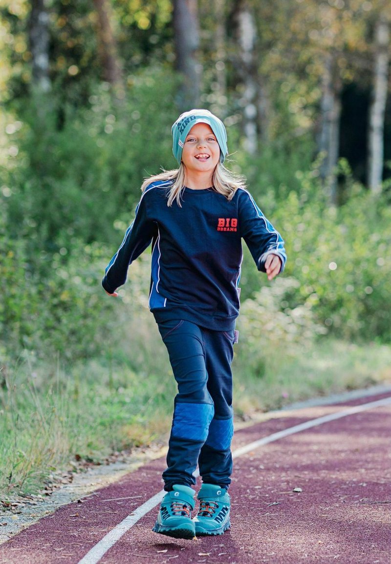 Tummansininen urheiluhuppari, jossa on punainen logo, yhdistettynä sinisiin housuihin, joissa on vaaleansinisiä yksityiskohtia. Päässä turkoosi pipo ja jaloissa saappaat.