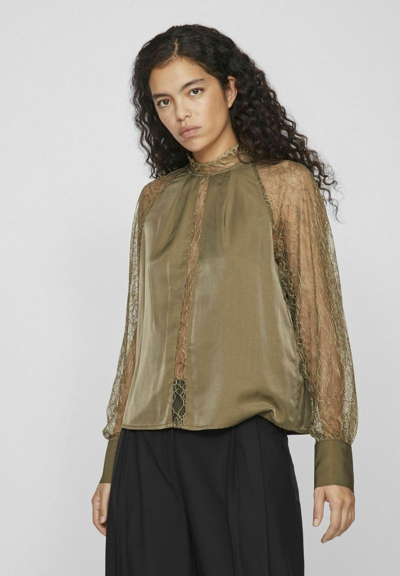 Blouse vert olive avec des manches longues en dentelle transparente, présentant un col haut et des plis subtils, complétée par un corps lisse et texturé.