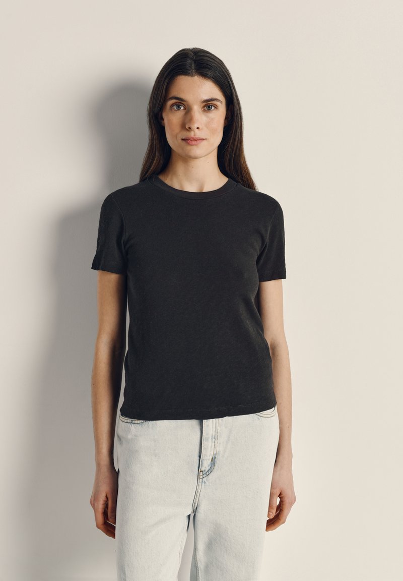 Jauna moteris su ilgais tamsiais plaukais dėvi juodą trumpomis rankovėmis marškinius ir šviesiai mėlynas džinsines kelnes, stovinti priešais paprastą baltai gelsvą sieną.