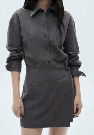 Kobieta ubrana w ciemnoszarą koszulę na guziki z długim rękawem podwiniętym do łokci oraz pasującą asymetryczną spódnicę na jednolitym tle.