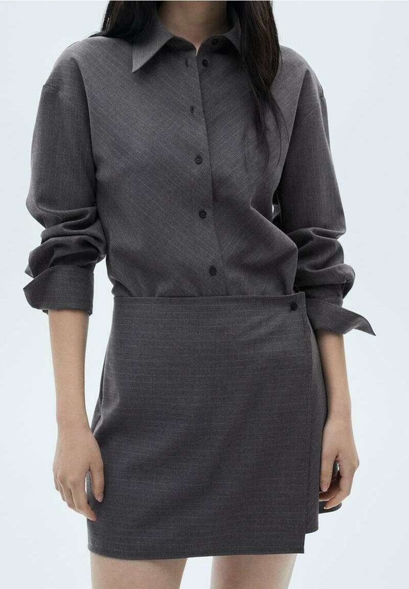 Femme portant une chemise boutonnée à manches longues gris foncé avec les manches retroussées et une jupe asymétrique assortie, sur un fond uni.