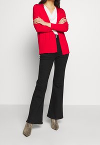 Rote offene Strickjacke über einem weißen V-Ausschnitt-Top, kombiniert mit schwarzen ausgestellt geschnittenen Hosen und beigen, stiefeletten mit Schlangenmotiv.