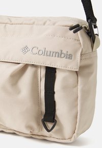 Beige stof skuldertaske med lynlås, med sort rem og hardware. Columbia-logoet prægede på frontklappen. Tekstureret overflade.