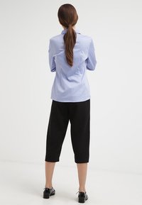 Camisa de botón de color azul claro con cuello y mangas largas, combinada con pantalones negros cortos y zapatos negros, vista desde la espalda.