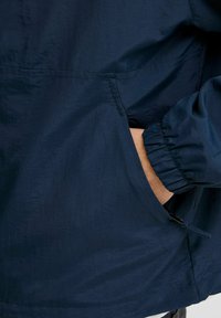 Veste légère bleu marine avec un tissu texturé, des poignets élastiques et une poche frontale. Présente un bas resserré avec un cordon de serrage.