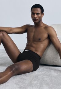 Boxer noir ajusté avec un logo crocodile Lacoste blanc sur la ceinture, porté par une personne à la peau mate, assise sur une surface claire.