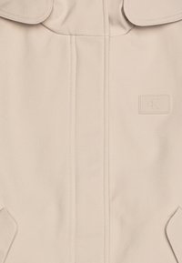 Beige jakke laget av glatt stoff, med logo-detalj, subtil søm, og et design med avrundede revers og sidelommer.