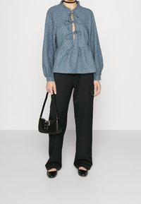 Blusa blu con texture, colletto alto, chiusura anteriore con fiocco e design peplum, abbinata a pantaloni neri a gamba larga e una piccola borsa nera.