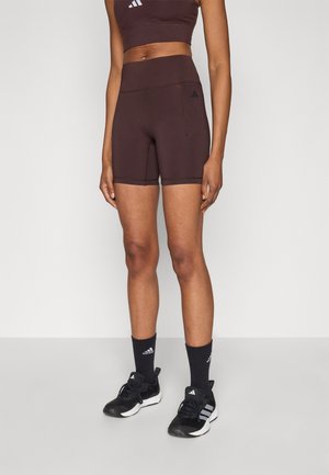 Brown athletic shorts made of stretchy material, high-waisted with a side pocket and subtle branding on the waistband, paired with black sneakers and socks.