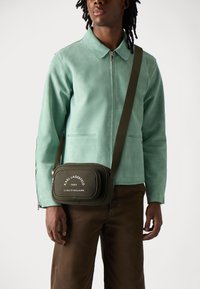 Giacca in suede verde con colletto, abbinata a pantaloni marroni. Piccola borsa oliva con testo bianco e tracolla regolabile.