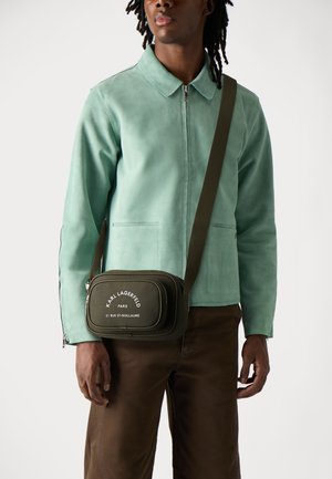 Grüne Wildlederjacke mit Kragen, kombiniert mit braunen Hosen. Kleine olivfarbene Tasche mit weißem Text und verstellbarem Gurt.