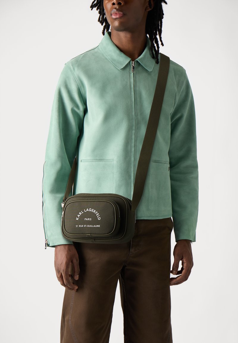 Giacca in suede verde con colletto, abbinata a pantaloni marroni. Piccola borsa oliva con testo bianco e tracolla regolabile.