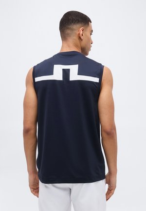 Homme portant un débardeur bleu marine avec un motif géométrique blanc sur le haut du dos, debout de dos sur un fond uni.