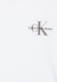Bijela pamučna majica sivi otisnuti logo "Calvin Klein Jeans" s istaknutim dizajnom "CK" na lijevoj strani prsnog područja.
