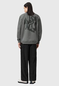 Felpa grigia con grande testo grafico nero sul retro, abbinata a pantaloni cargo neri e sneaker bianche. Stile casual, vestibilità rilassata.