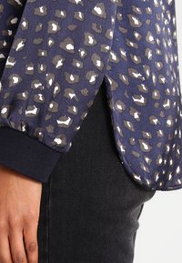 Blusa navy con un motivo leopardato in tonalità di marrone e bianco. Presenta un polsino a coste e spacco laterale. Il tessuto appare liscio e leggero.