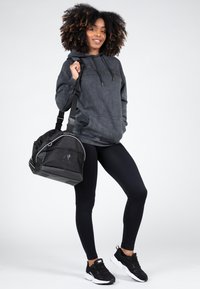 Zwarte sporttas met netpaneel, zilveren accenten en logo. De drager draagt een grijze hoodie en zwarte leggings, gecombineerd met zwarte sportschoenen.