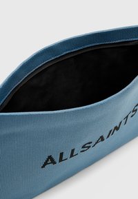 Sacco in tessuto blu chiaro con superficie texturizzata, caratterizzato dalla scritta nera "ALLSAINTS" lungo il lato e un interno contrastante di colore nero.