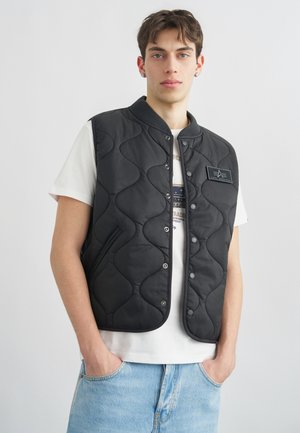 Jonge man met kort bruin haar, gekleed in een zwart doorgestikt vest over een wit t-shirt en lichtblauwe spijkerbroek, handen in de zakken, neutrale achtergrond.