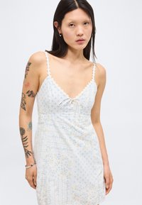 Νεαρή γυναίκα με μακριά σκούρα μαλλιά που φοράει λευκό αμάνικο φόρεμα με floral μοτίβο και δαντελωτούς ιμάντες, δείχνοντας τατουάζ στο αριστερό της χέρι.