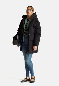 Černá péřová bunda s kapucí, se zipem vpředu a elastickými manžetami; nošena přes proužkované triko a kombinována s modrými džíny a černými balerínami.
