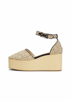 Beiger Plateau-Keilabsatz-Schuh mit schwarzem geometrischem Muster, Knöchelriemen mit goldener Schnalle und geschlossenem spitz zulaufendem Vorderteil auf einfarbig hellem Hintergrund.